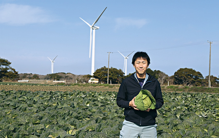 新品種「結岬」を手にするJAちばみどりの中島慎平さん。風力発電の風車が、風の強さを物語っている。